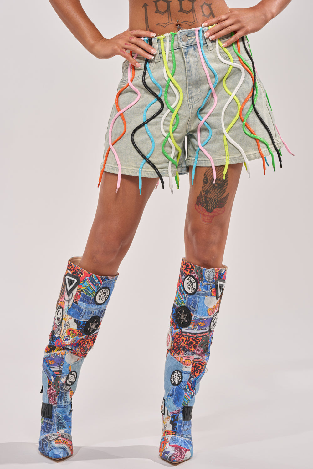 A person wears the CRAZY FOR YOU DENIM SHORT with rainbow laces and colorful wavy cords at the hem, paired with tall graphic patchwork boots. Hands on hips reveal torso and thigh tattoos—a bold festival streetwear vibe. 2