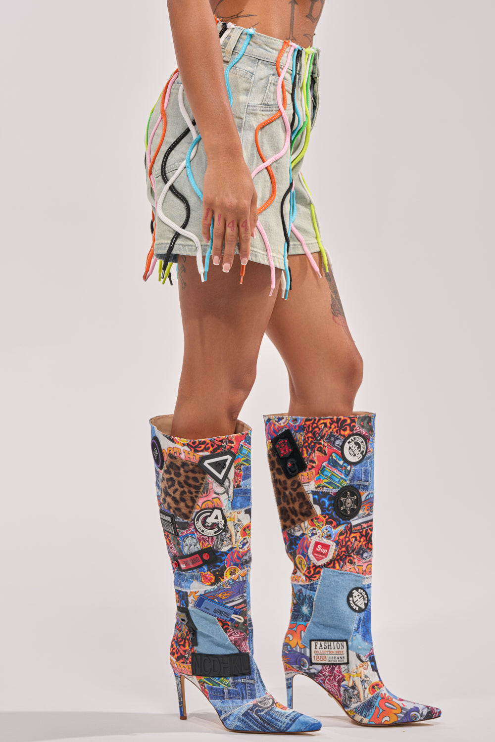 Someone stands sideways in the CRAZY FOR YOU DENIM SHORT, featuring rainbow lace and colorful wavy trim, paired with tall, pointed-toe boots with vibrant graphic patches—an eye-catching streetwear look against a plain light background. 3