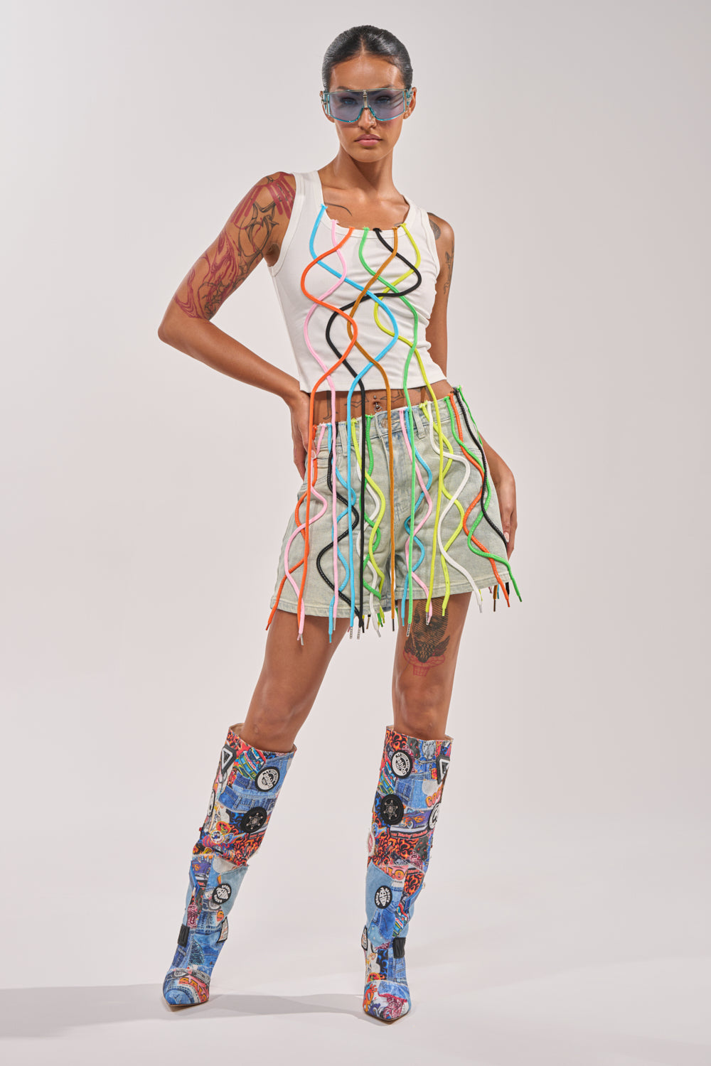 A model wears sunglasses, a white tank top with colorful wavy lines, the CRAZY FOR YOU DENIM SHORT with hanging multicolored strings, and knee-high boots with patchwork cartoon patterns, standing against a plain background. 6