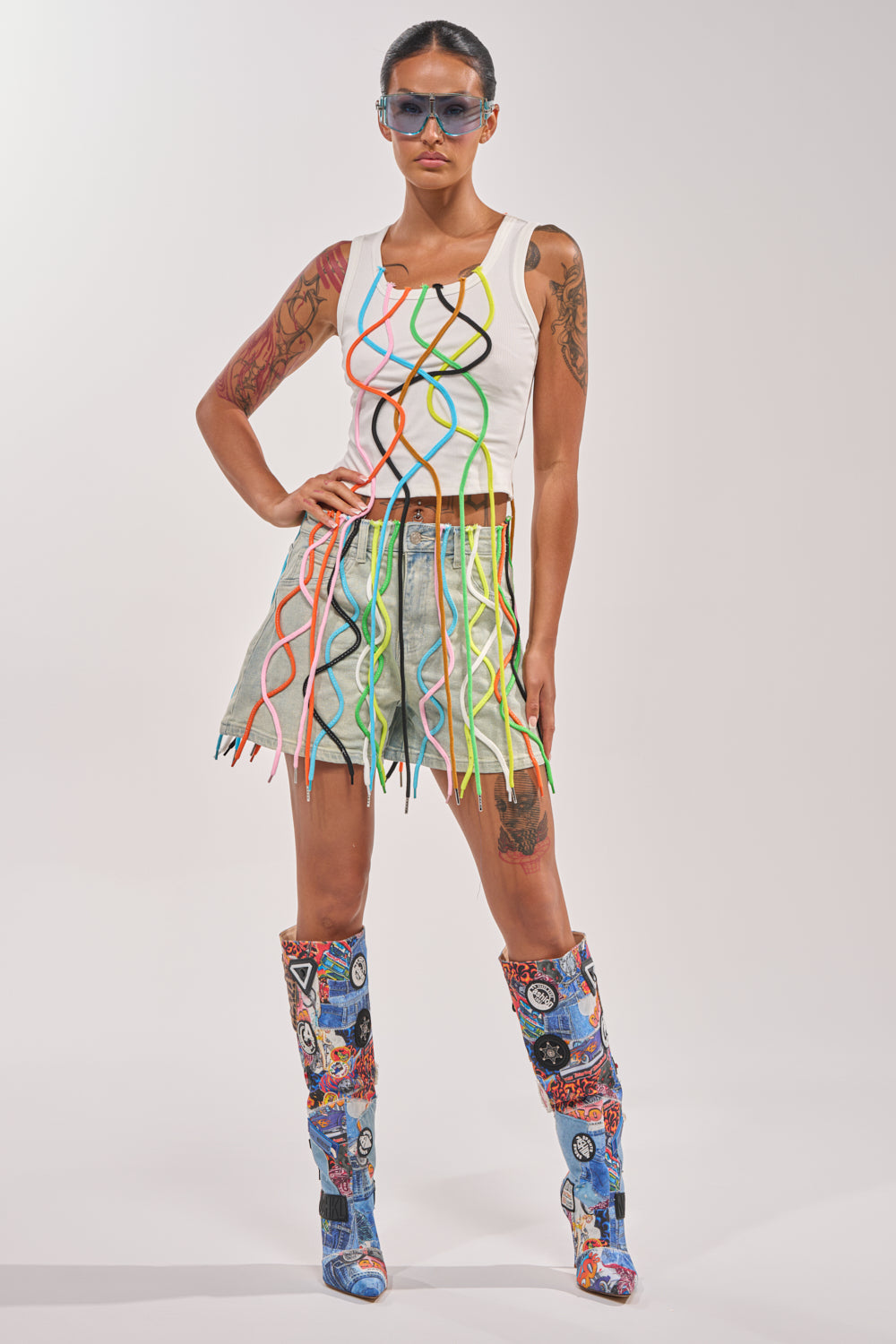 A woman poses in a white tank top, CRAZY FOR YOU DENIM SHORT, patterned knee-high boots, and blue-tinted sunglasses—an eye-catching look perfect for festival streetwear. 7