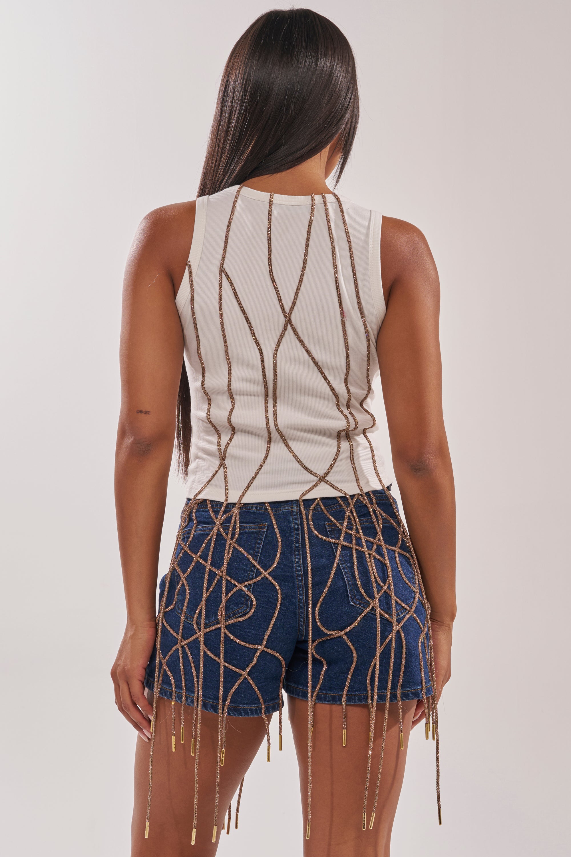 A person with long, straight hair wears the NINA EMBELLISHED RIBBED TANK and denim shorts. Gold decorative chains and embellished fringe form a web-like pattern cascading down the back and legs of the outfit. 5