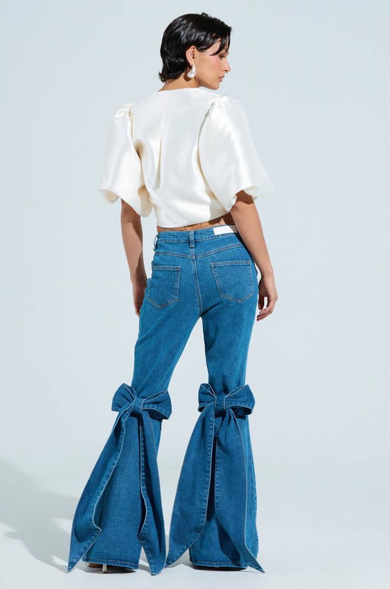 Facing away in a plain light blue backdrop, a person wears ALL TIED UP BOW FLARE JEANS paired with a cream blouse. 1