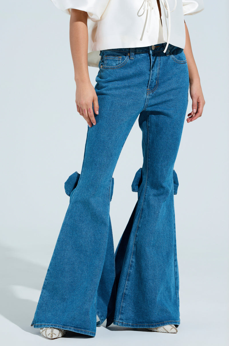 A person in ALL TIED UP BOW FLARE JEANS, featuring high-rise and flared design with large side bows, paired with a white blouse. They wear textured white shoes against a plain, light background. 2