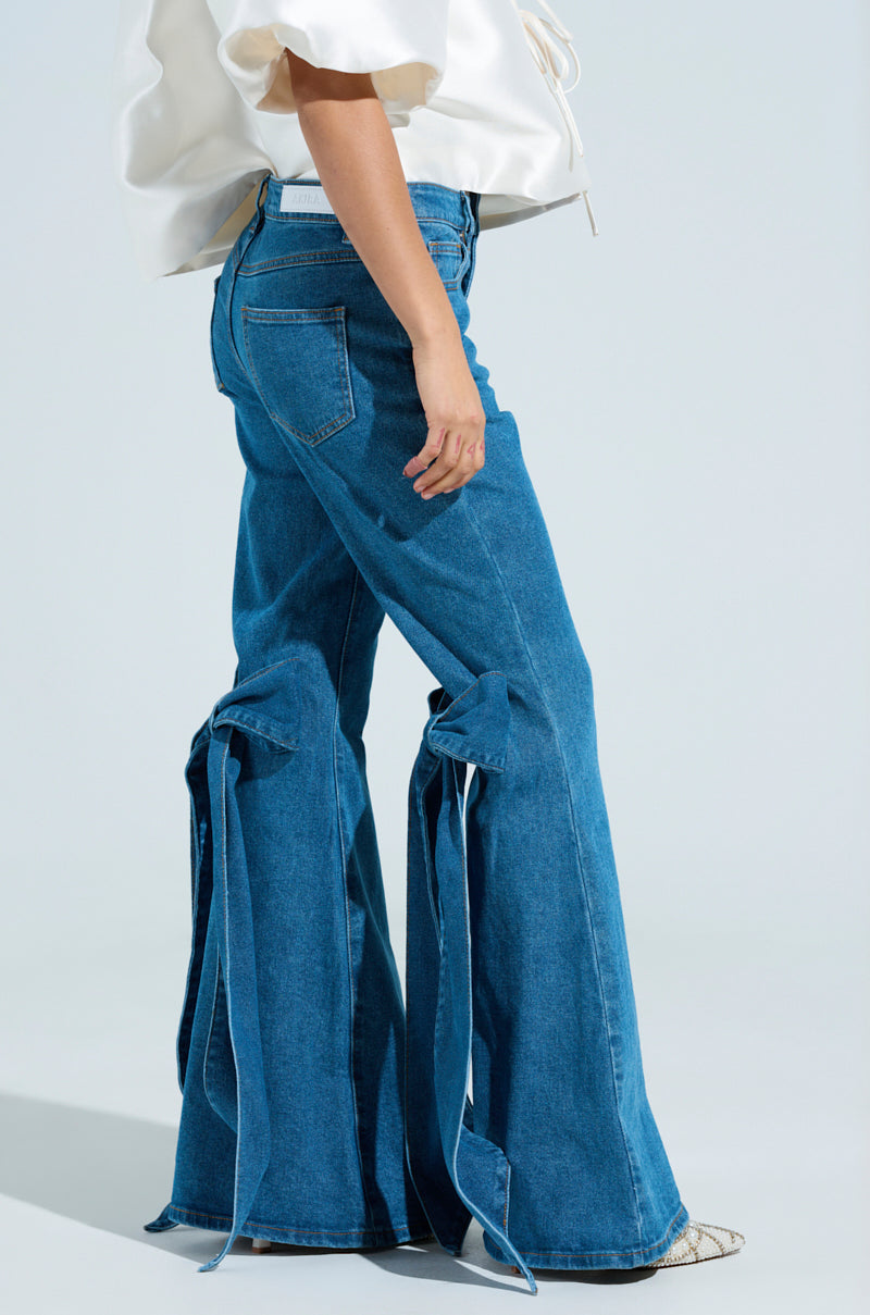 Wearing ALL TIED UP BOW FLARE JEANS, featuring high-rise fit and large bows near the ankles, paired with a white top and classic pocket design. Pointy textured shoes complete the look against a plain light gray background. 3