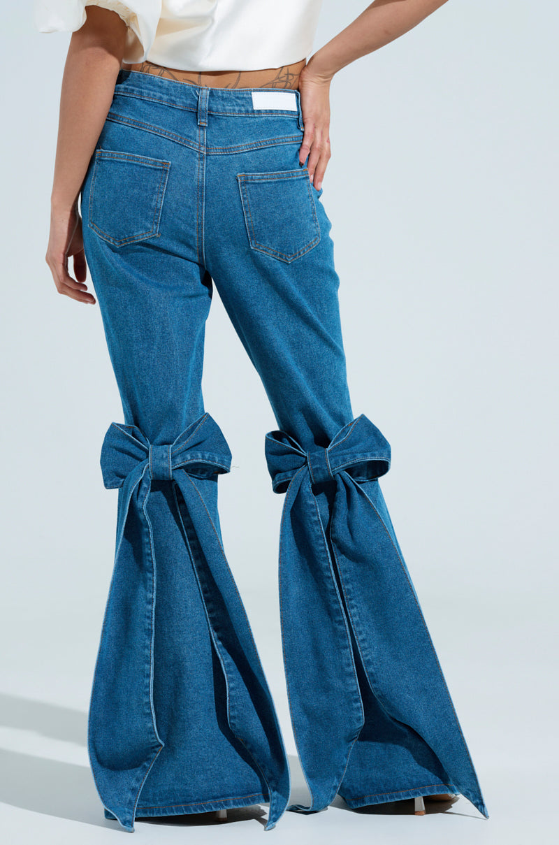 A person wearing ALL TIED UP BOW FLARE JEANS stands confidently, one hand on their hip. The jeans showcase large bows on each flared leg, prominent back pockets, and a white label on the waistband. 0