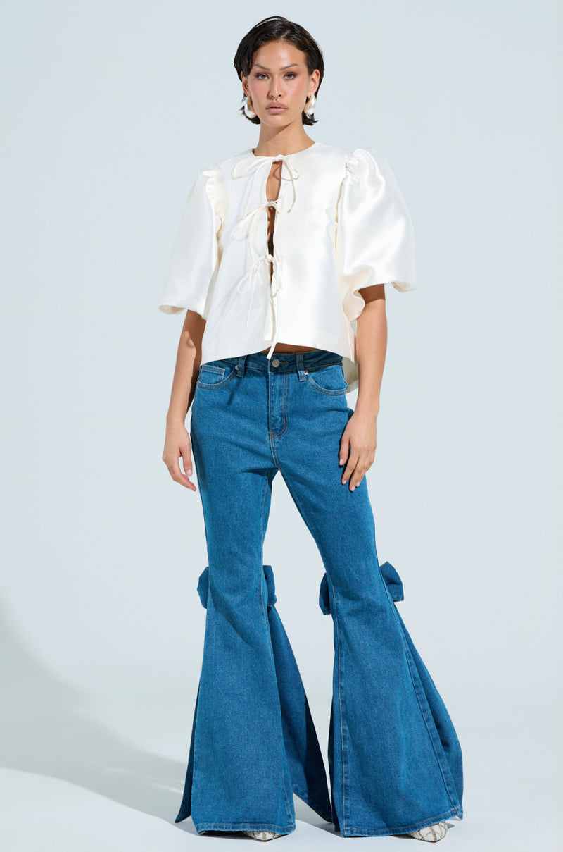 A person exudes confidence in a white blouse with puffed sleeves and ALL TIED UP BOW FLARE JEANS. With short hair and earrings, they pose against a plain backdrop. 4