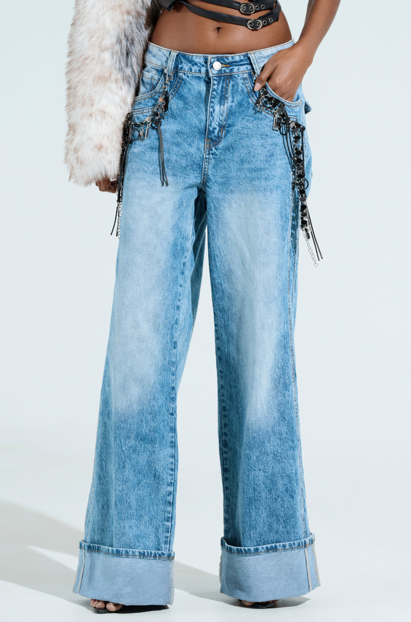 Someone is wearing the ZIP ZIP ZIPPER CHARM JEANS, featuring high-rise, wide-leg denim in light blue with embellished chains and tassels on the pockets. The jeans also have a fur-like sleeve and folded hems, set against a plain light backdrop for a striking effect.