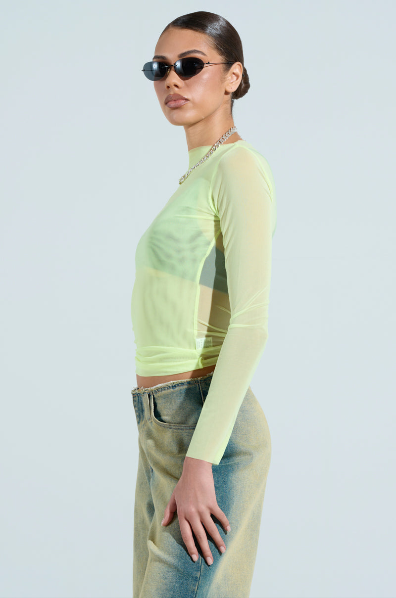 A person wearing the Paxton Sexy Double Lined Mesh Long Sleeve Top in light green, together with faded jeans, sunglasses, and a necklace, stands in profile against a neutral background.