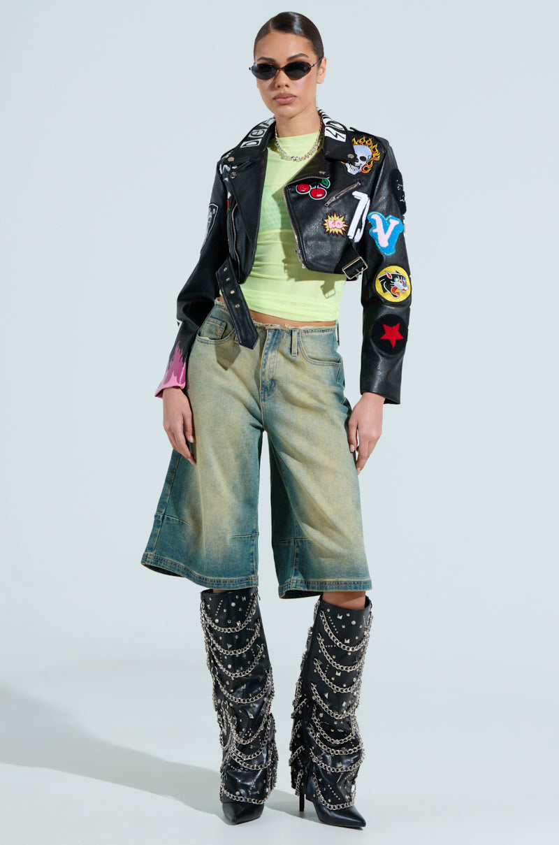 Wearing sunglasses and a black leather jacket with colorful patches, someone rocks the Paxton Sexy Double Lined Mesh Long Sleeve Top, denim culottes, and knee-high black boots with chain details. This club-ready outfit exudes confidence against a plain background.