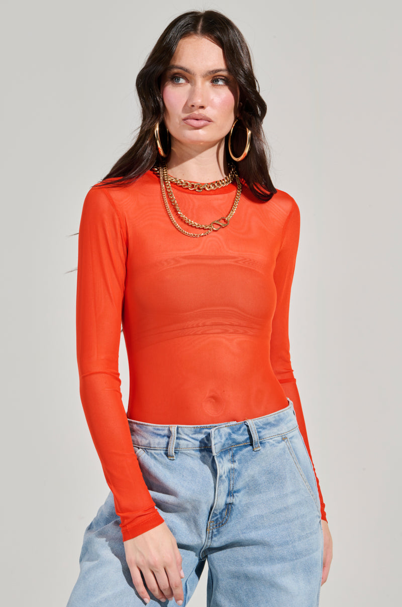 A person with long dark hair wears the Paxton Mesh Long Sleeve Crew Neck Bodysuit in orange, paired with light blue jeans. They accessorize with large hoop earrings and layered gold necklaces against a plain gray background.