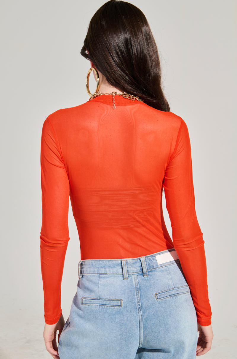 A person with long, dark hair stands with their back to the camera, wearing the PAXTON MESH LONG SLEEVE CREW NECK BODYSUIT IN ORANGE paired with light blue jeans featuring a visible white label. Large hoop earrings and a gold necklace complete the curve-enhancing ensemble.