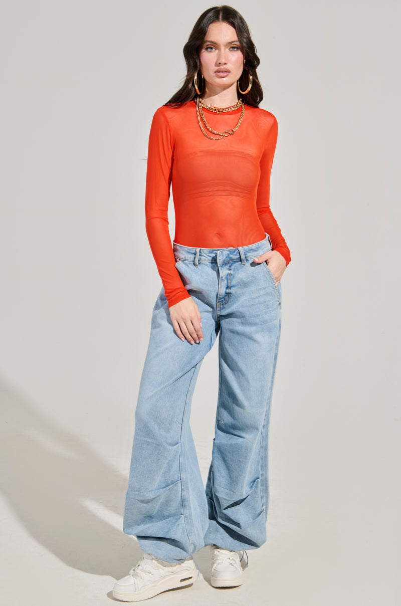 A person stands against a plain background wearing the PAXTON Mesh Long Sleeve Crew Neck Bodysuit in Orange, paired with light blue wide-leg jeans, white sneakers, and gold hoop earrings. Their curve-hugging fit complements their long dark hair as they casually keep their hands in pockets.
