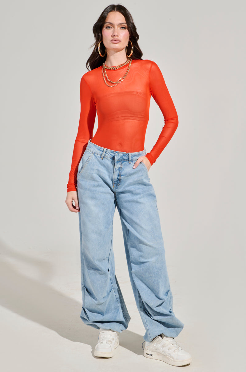 A person wearing the PAXTON Mesh Long Sleeve Crew Neck Bodysuit in Orange, which is sheer and curve-hugging, is paired with baggy light blue jeans and white sneakers. Accessories like large hoop earrings and layered necklaces complement the outfit as they stand against a neutral background.