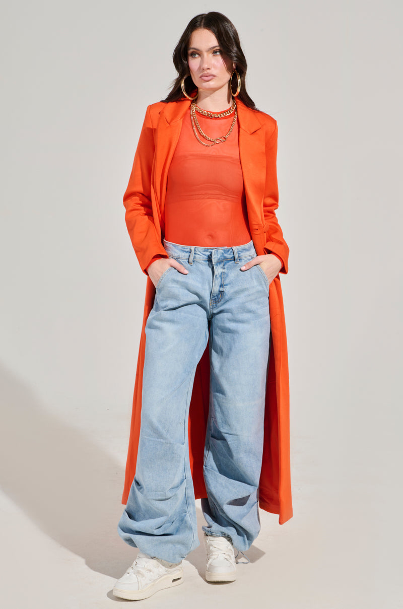 A person confidently poses in the PAXTON MESH LONG SLEEVE CREW NECK BODYSUIT IN ORANGE, paired with an orange long coat. They have their hands tucked into the pockets of their super stretchy, curve-hugging light blue jeans and are wearing white sneakers, a gold necklace, and hoop earrings against a neutral background.