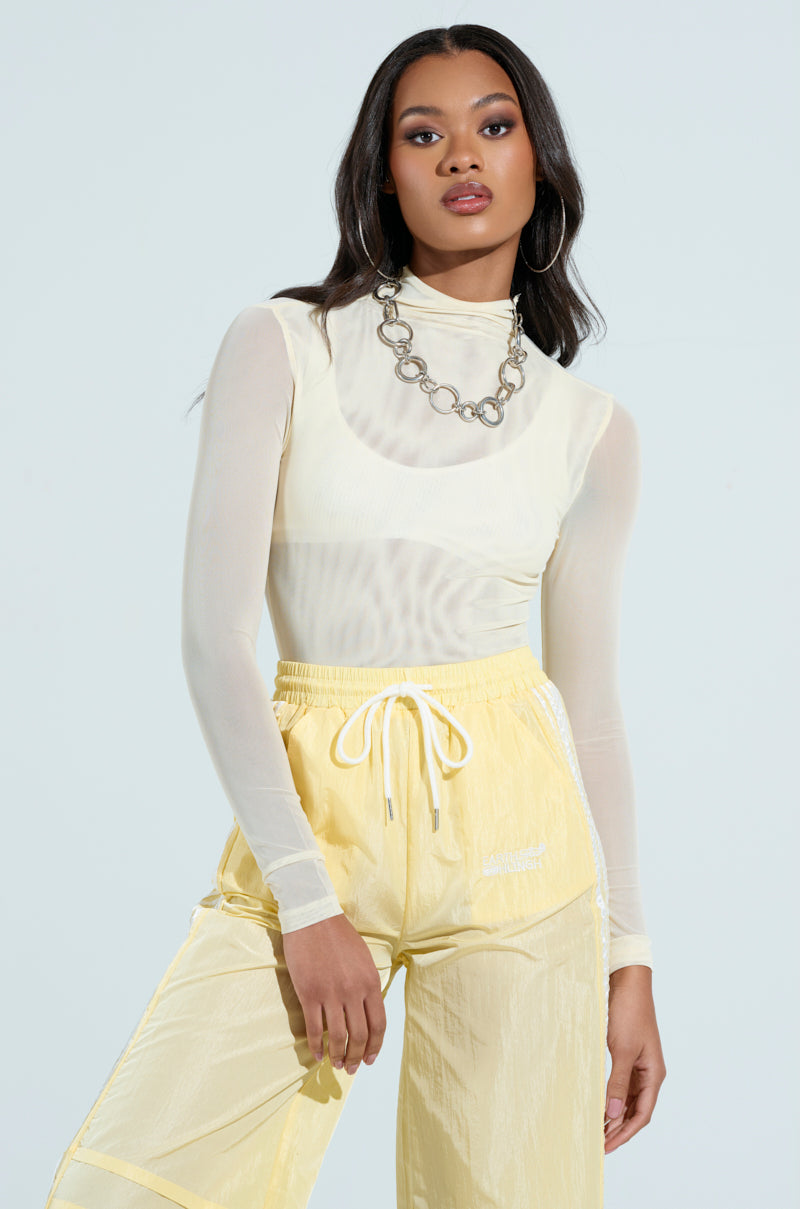 The woman strikes a pose in a PAXTON SEXY MESH LONG SLEEVE MOCKNECK BODYSUIT and light yellow pants, accessorized with a chunky chain necklace. Her long, wavy hair complements her festival look against the light background.