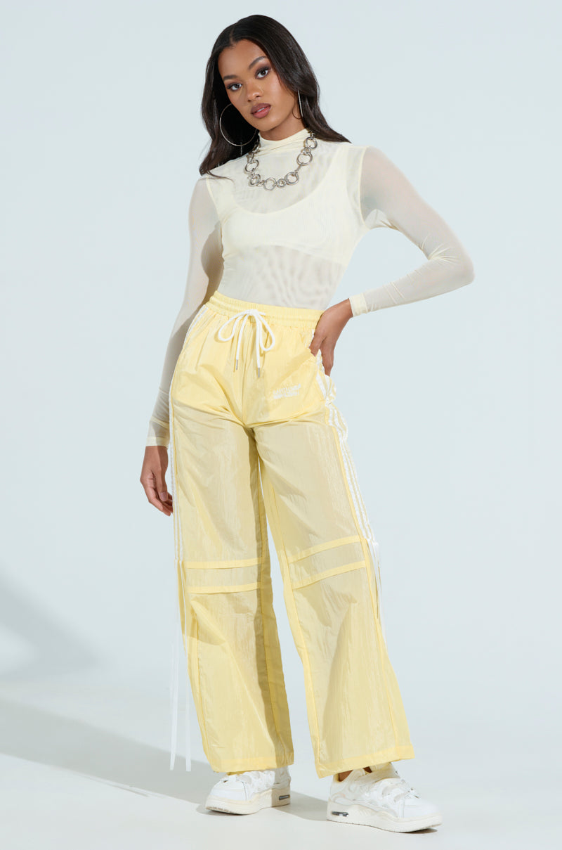 A woman confidently wears the Paxton Sexy Mesh Long Sleeve Mockneck Bodysuit paired with a silver necklace and yellow drawstring pants. She completes her festival look with white sneakers and hoop earrings against a plain backdrop.