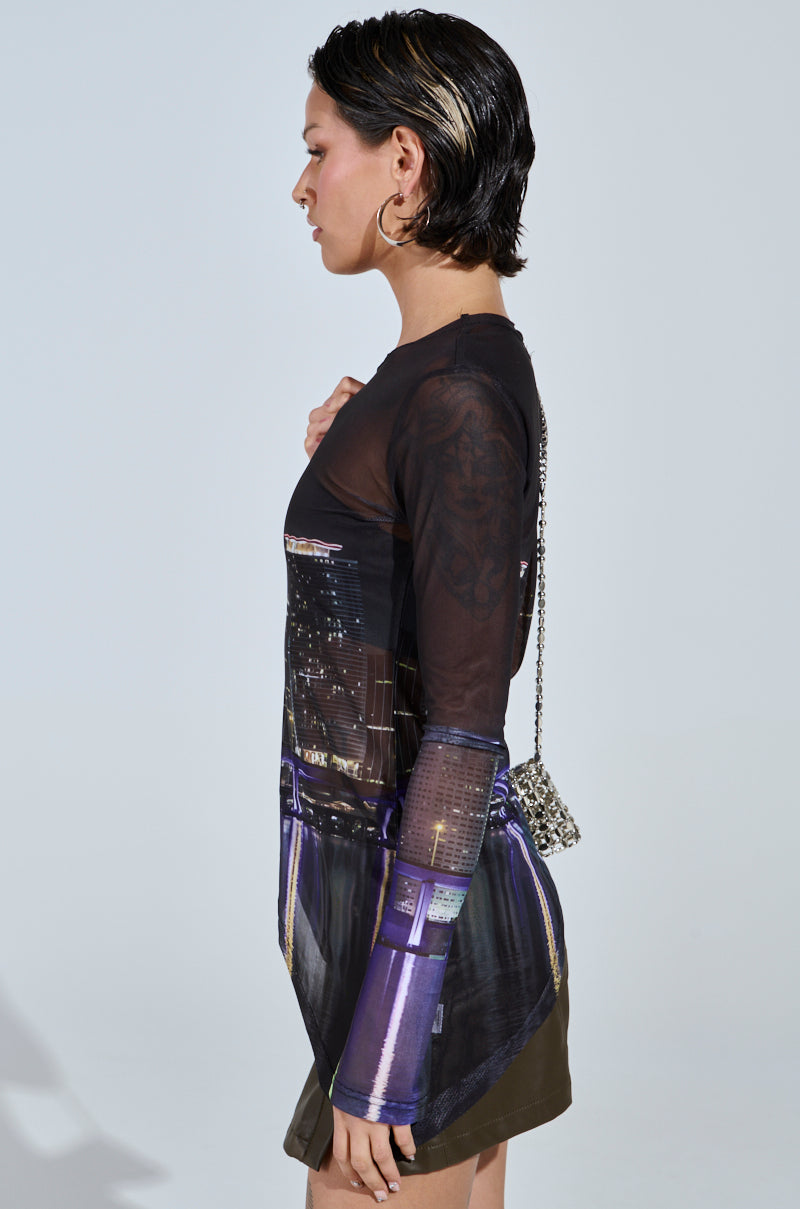 A person with short dark hair wears the CITY LIFE MESH TOP, featuring a cityscape graphic, and hoop earrings. They hold a small chain-strap bag against a plain backdrop, maintaining a neutral expression.