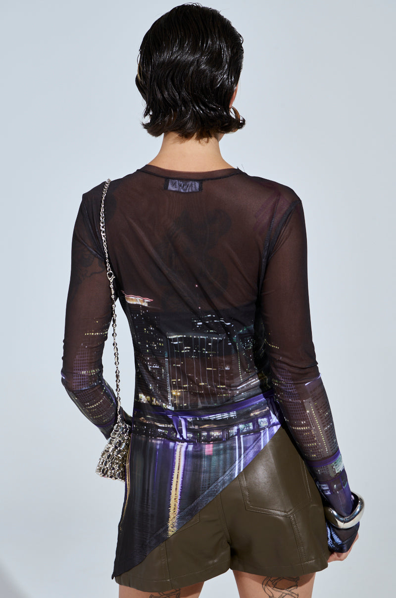 A person with short, dark hair is seen from behind, wearing the CITY LIFE MESH TOP with a cityscape graphic and dark shorts. They have a small chain-strap bag over one shoulder against a plain light gray background.