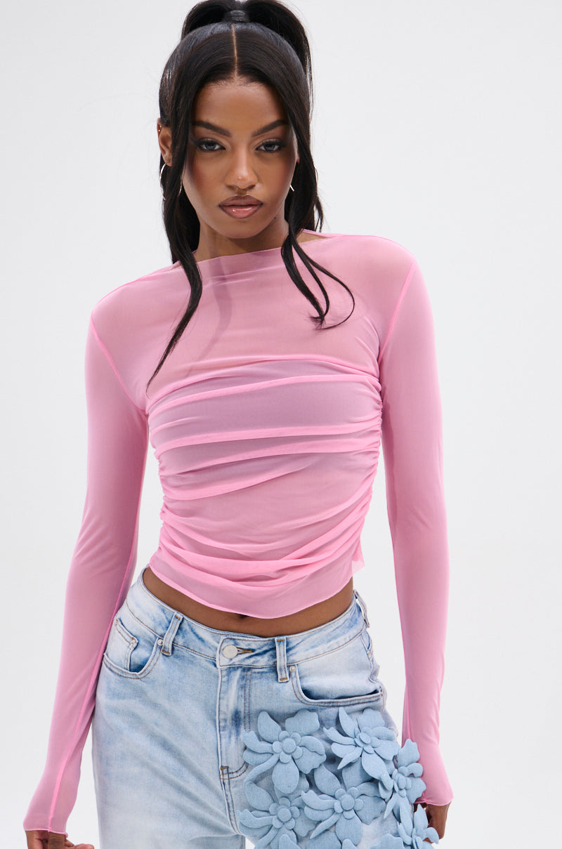 Wearing the PAXTON Sexy Ruched Mesh Long Sleeve Top in pink, a woman pairs it with high-waisted light blue jeans featuring large fabric flowers. Her hair is styled in high ponytails as she poses against a neutral backdrop, highlighting her sculpted silhouette.