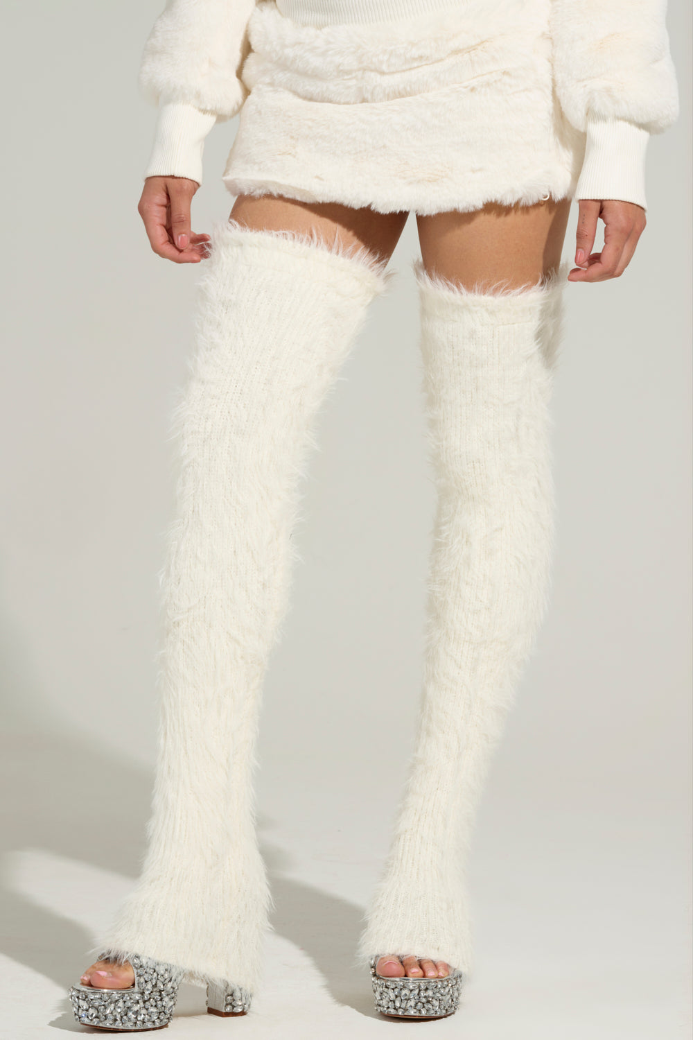 A person stands against a plain background wearing a fuzzy white outfit that includes a mini skirt, matching WARM AND COZY THIGH WARMERS, and chunky high-heeled shoes with sparkling details. Their ensemble showcases faux mohair textures and stretchy knit fabrics, adding an extra layer of cozy sophistication.