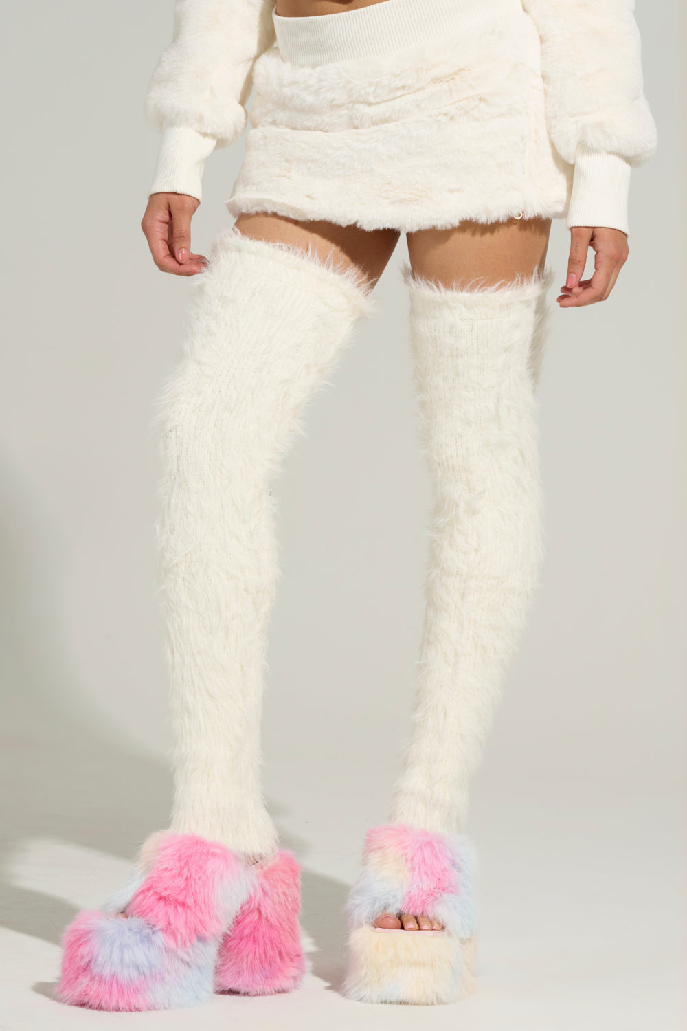 A person showcases a fuzzy white two-piece ensemble consisting of a cropped top and mini skirt. They complete the look with WARM AND COZY THIGH WARMERS, featuring faux mohair material and elasticized bands, paired with colorful fluffy platform shoes in pink, blue, and white hues. The plain background ensures all attention remains on this stunning outfit. 1