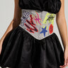 A person wearing a black strapless dress with a vibrant, artistic corset showcasing graffiti-inspired designs such as flames and stars. The dress features a bubble skirt, cinched with the PATCHED UP STRETCH DENIM BELT. They finish off the look with a silver necklace adorned with a cross pendant.