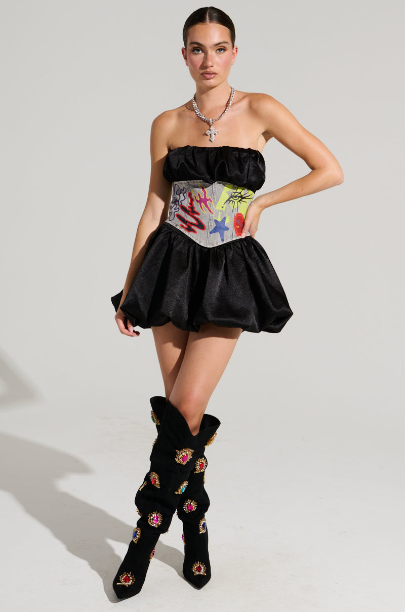 A person poses in a short, strapless black dress paired with matching high-heeled boots. The outfit is accented by the PATCHED UP STRETCH DENIM BELT, which boasts colorful, graffiti-inspired patterns in red, blue, and yellow. They stand against a plain light background. 1