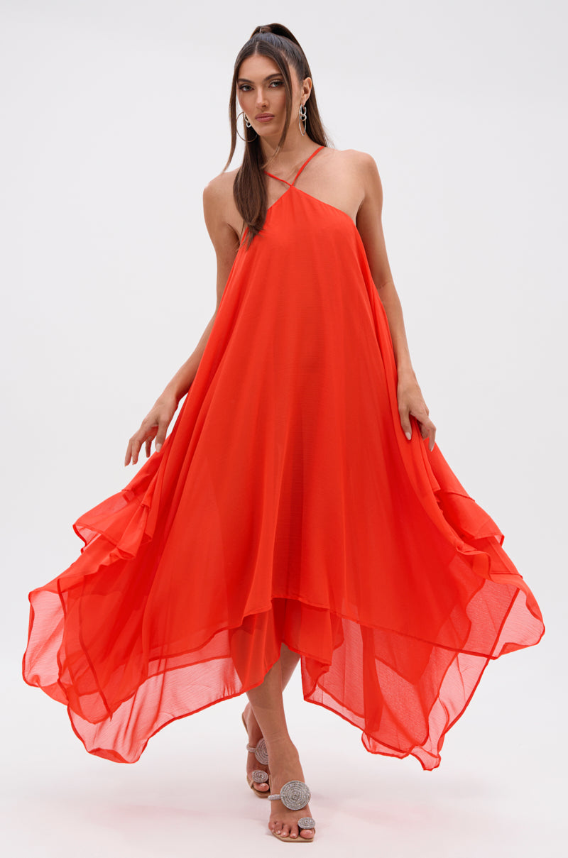 A woman models the RUNAWAY CHIFFON MAXI DRESS, featuring a flowing bright red chiffon fabric, asymmetrical hem, and halter neckline. She pairs it with silver open-toe sandals and hoop earrings against a simple light background.