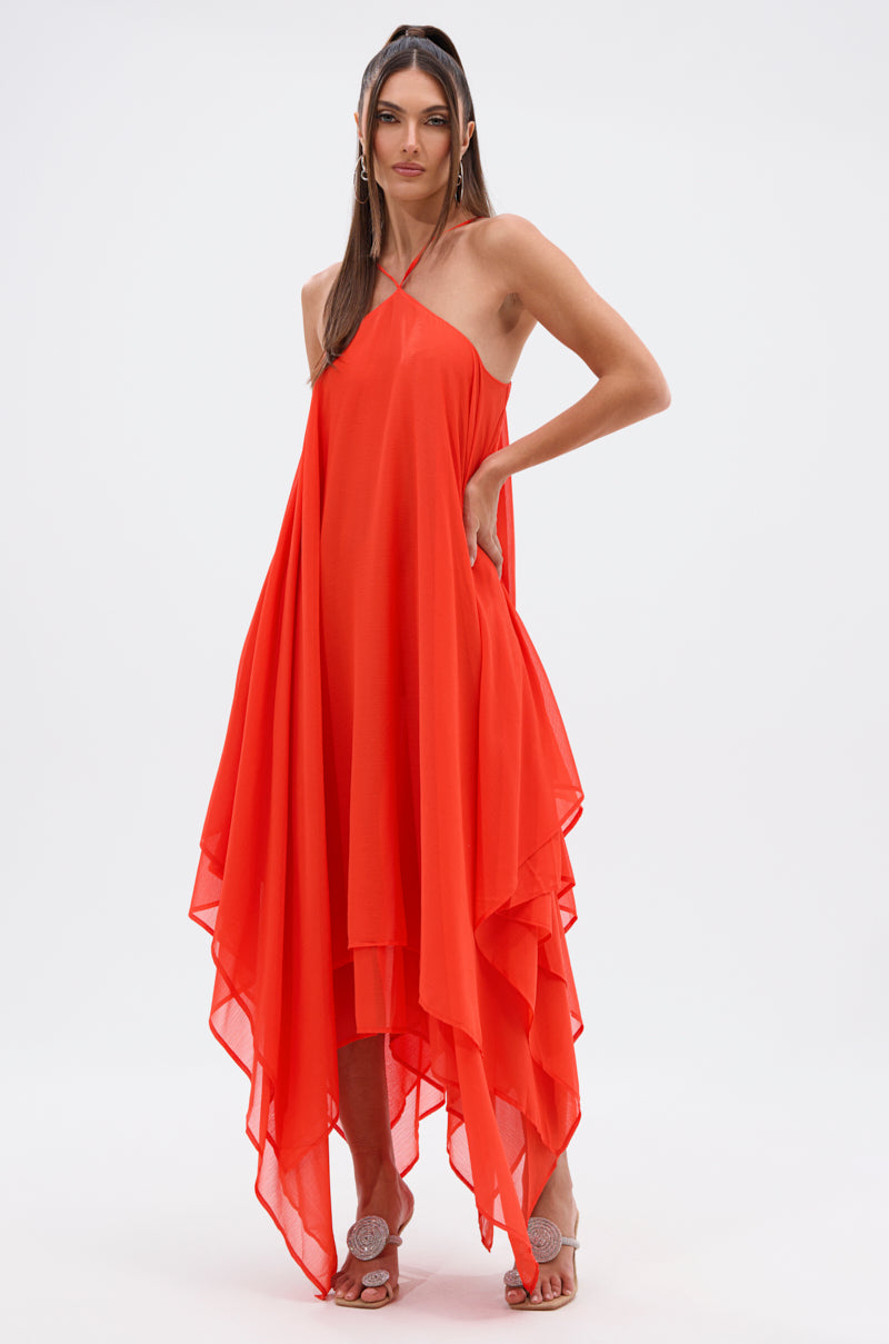 A woman with long straight hair poses against a plain white background, wearing the RUNAWAY CHIFFON MAXI DRESS—a flowing bright red maxi with thin straps and an asymmetrical hem—paired with white open-toe heeled sandals.