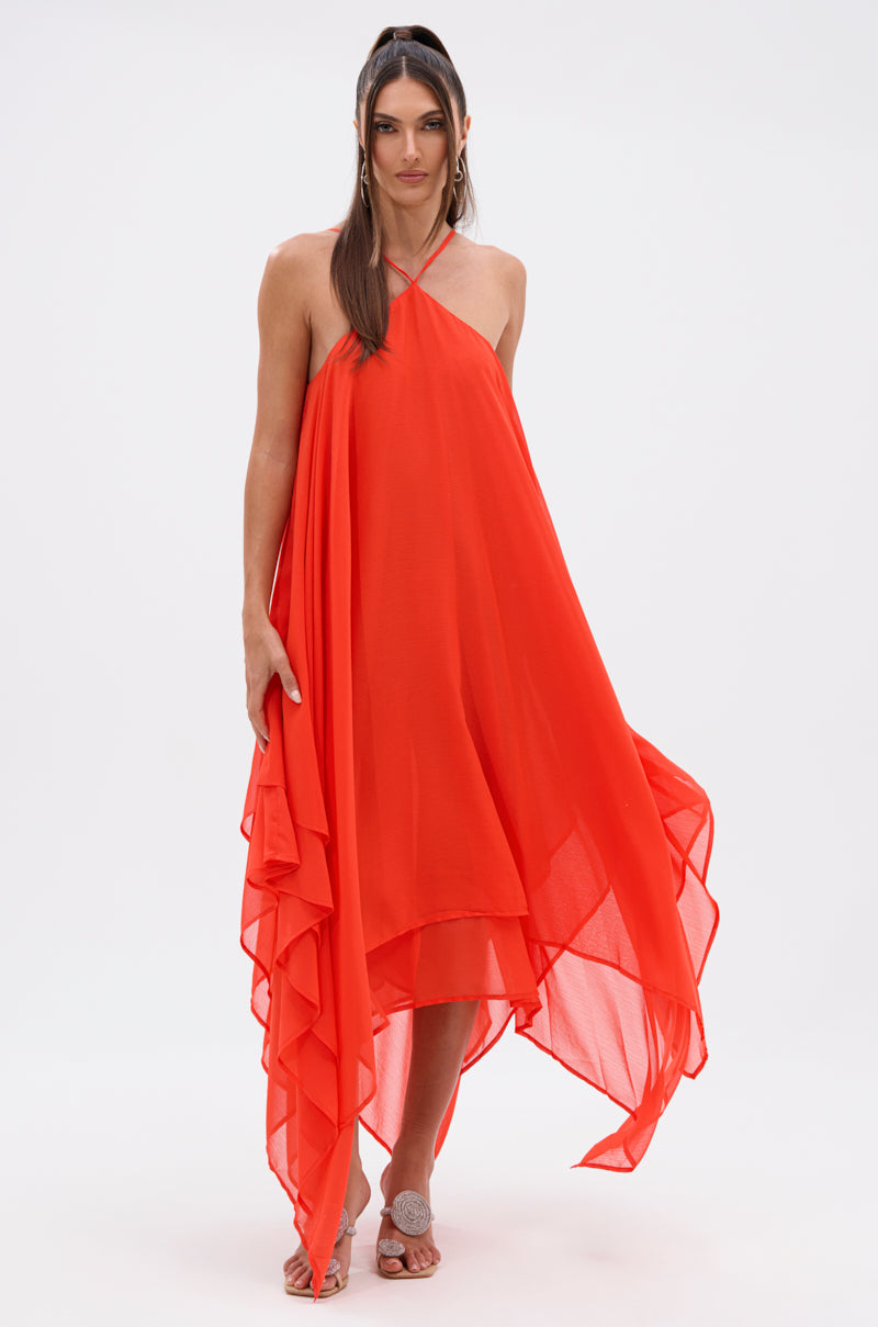 A woman wears the RUNAWAY CHIFFON MAXI DRESS in bright orange, featuring a halter neckline and an asymmetrical layered hem. She has long brown hair styled half-up, half-down and open-toed shoes against a plain white background.