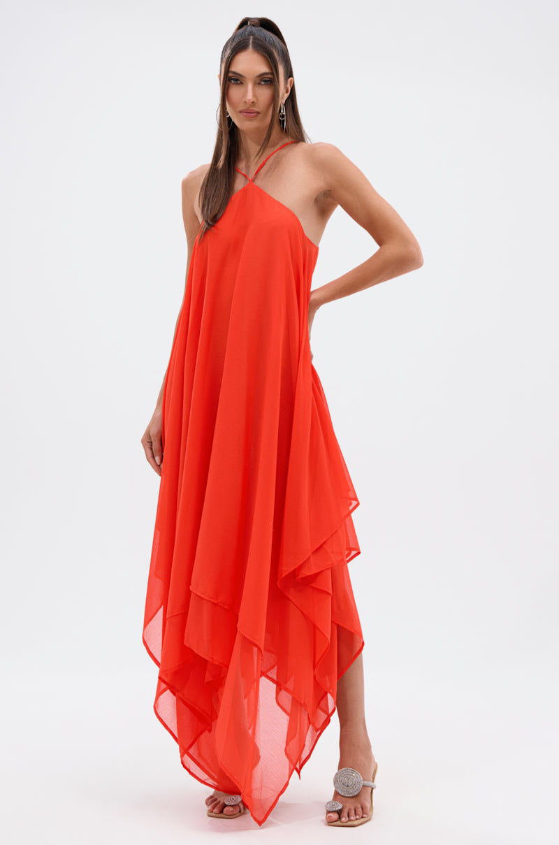 A woman stands confidently in the RUNAWAY CHIFFON MAXI DRESS, a bright orange maxi with thin straps and an asymmetrical handkerchief hem. She pairs it with hoop earrings and neutral heels against a plain white background.