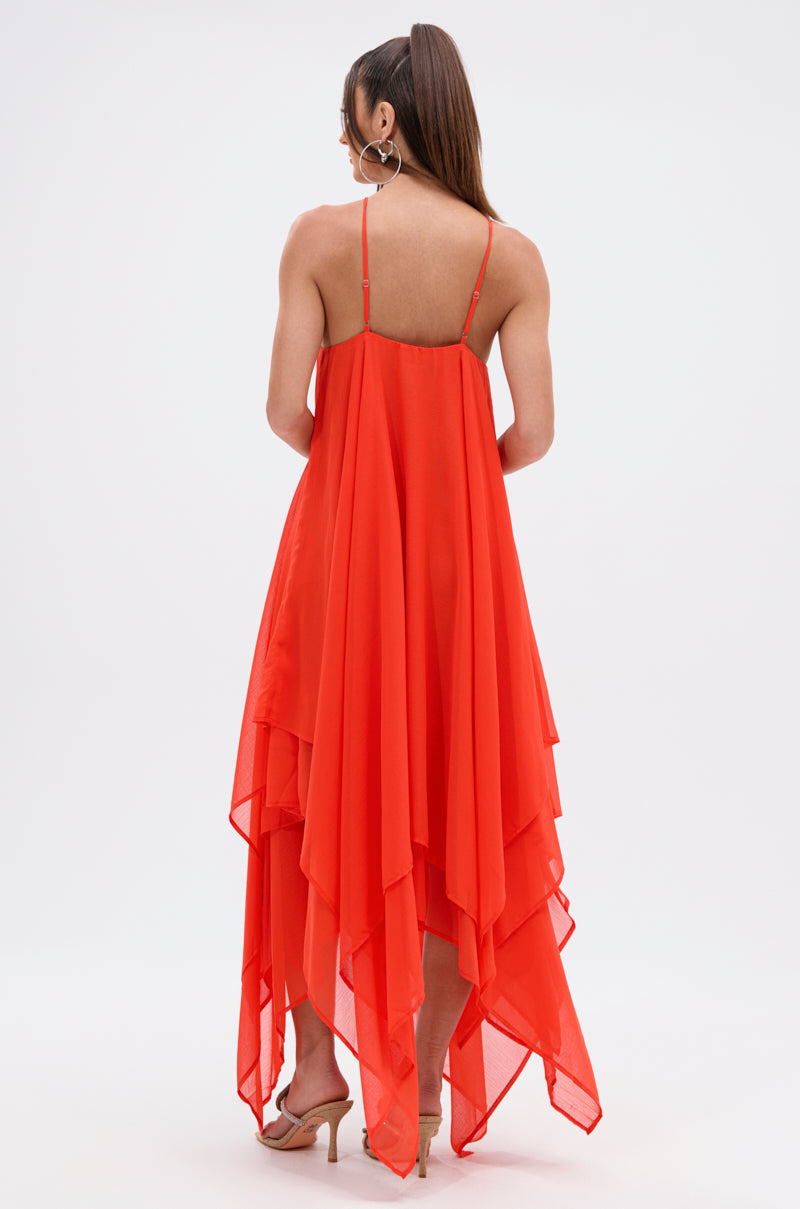 A woman with long hair in a ponytail models the RUNAWAY CHIFFON MAXI DRESS, featuring a vibrant orange hue, thin straps, and an asymmetrical hem. She stands facing away, highlighting the dress’s back and beige heels against a plain backdrop.