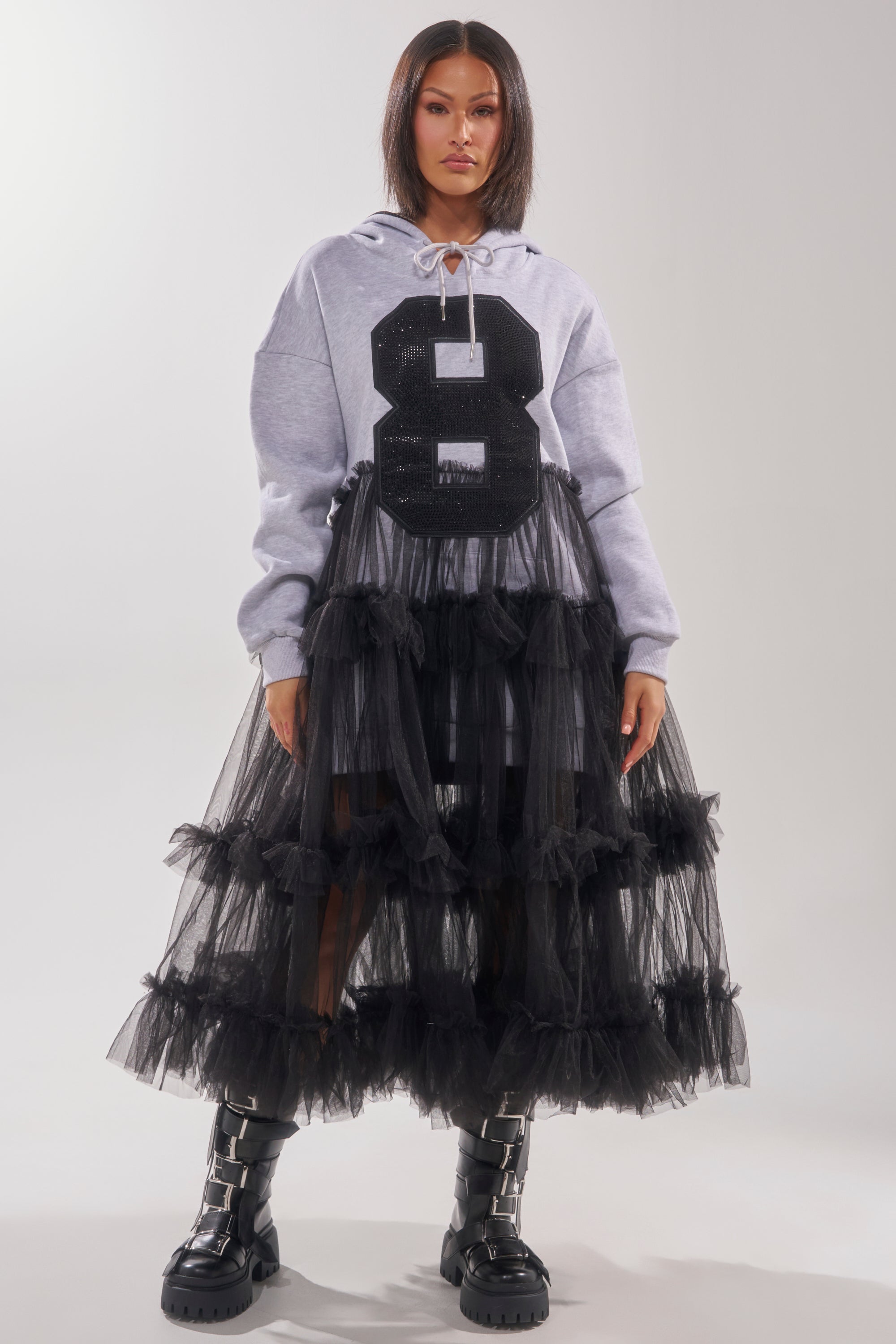 A woman exudes confidence in the DO SOMETHING CRAZY SWEATSHIRT DRESS, featuring a bold black "8," layered under a long black ruffle skirt and paired with chunky boots, set against a simple light backdrop.