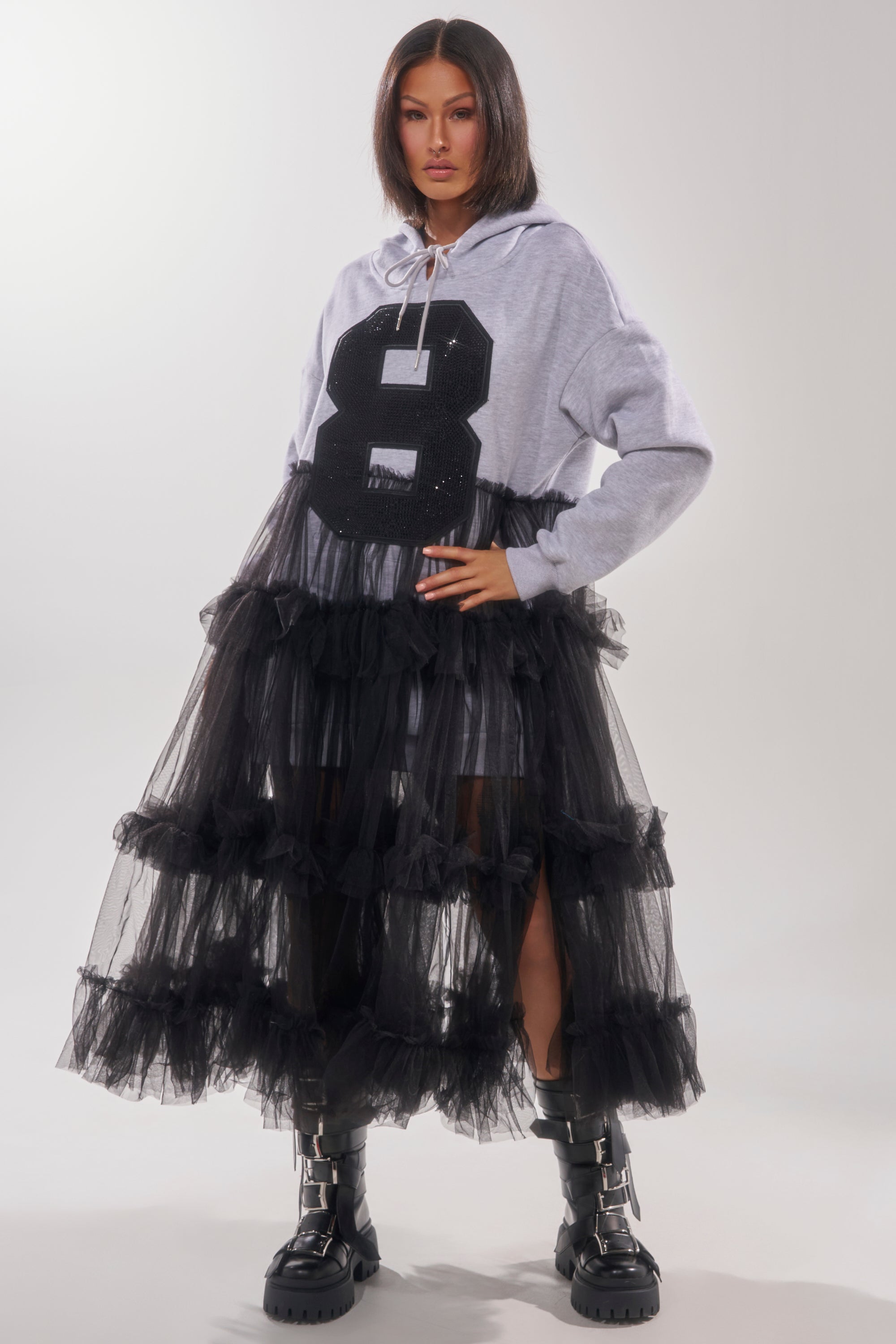 A woman exudes confidence in the DO SOMETHING CRAZY SWEATSHIRT DRESS, featuring a bold black "8" rhinestone varsity graphic, styled under a black tiered tulle skirt with black boots. She has straight, shoulder-length hair.