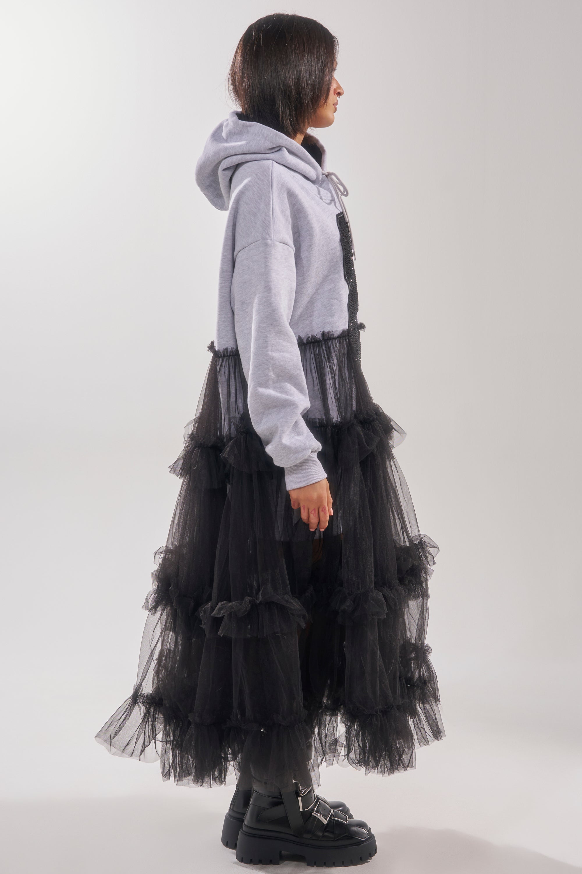 A person stands sideways wearing the DO SOMETHING CRAZY SWEATSHIRT DRESS layered with a long black ruffled tulle skirt and chunky black boots against a plain white background.
