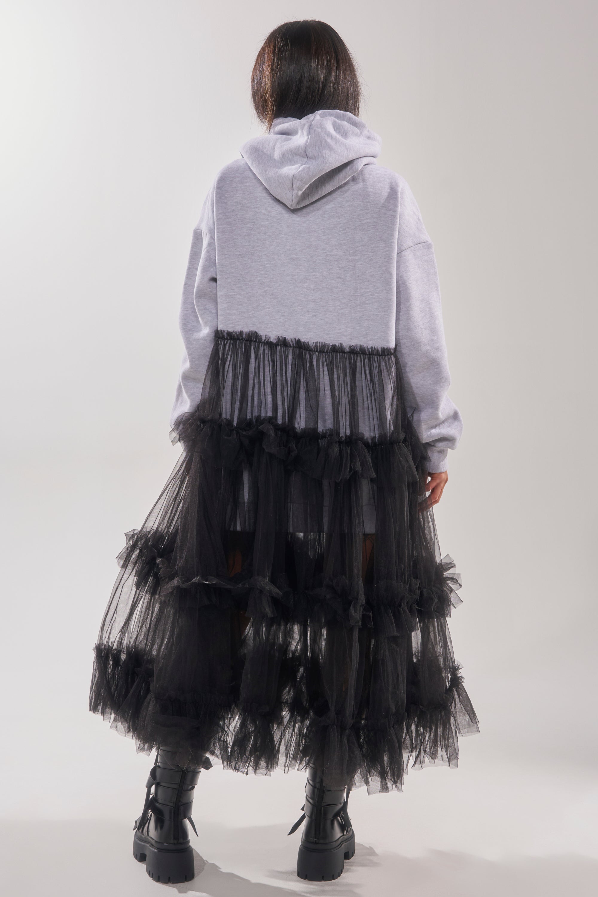 A person faces away, wearing the DO SOMETHING CRAZY SWEATSHIRT DRESS—a gray hoodie with ruffled black tulle layers from the waist down, resembling a dramatic maxi skirt—paired with chunky black boots.