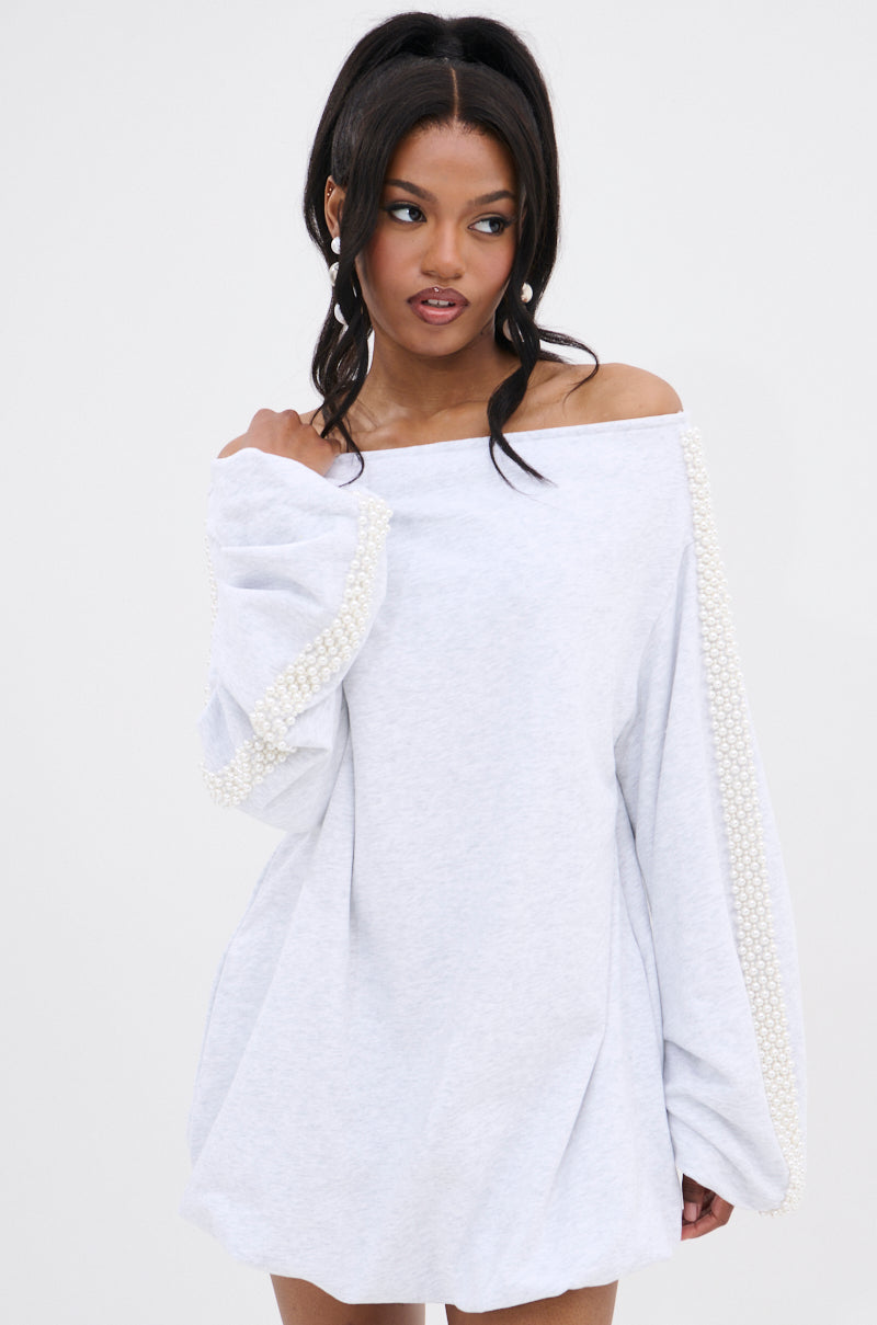 A woman with long dark hair in a high ponytail wears the GORGINA OFF THE SHOULDER SWEATSHIRT DRESS, featuring pearl-embellished sleeves, while looking to the side with a neutral expression against a plain background.