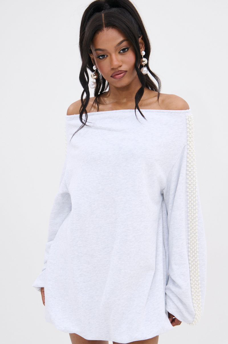 A woman with long dark hair in a high ponytail wears the GORGINA OFF THE SHOULDER SWEATSHIRT DRESS, featuring pearl embellishments on the sleeves. She accessorizes with large pearl earrings and stands against a plain light background.