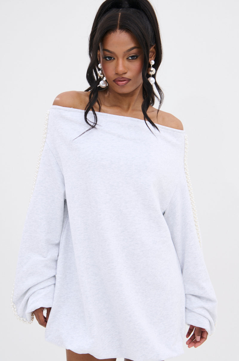 A woman wears the GORGINA OFF THE SHOULDER SWEATSHIRT DRESS, featuring a loose fit and pearl-embellished sleeves, styled with large pearl earrings and a high ponytail against a plain white background.