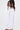 A woman with long, wavy dark hair models the GORGINA OFF THE SHOULDER SWEATSHIRT DRESS, featuring pearl-embellished sleeves. She stands sideways, looking ahead, and accessorizes with large dangling pearl earrings.