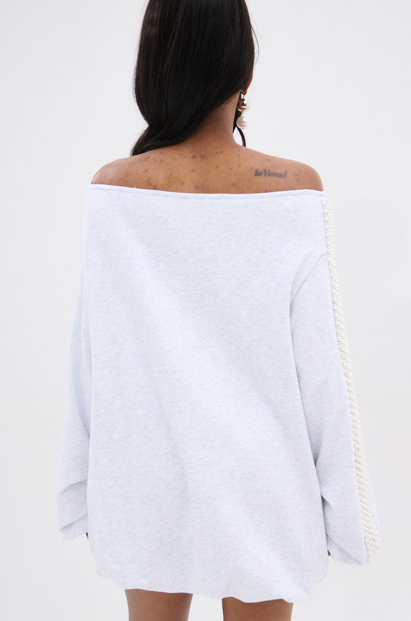 A woman with long dark hair faces away from the camera, wearing the GORGINA OFF THE SHOULDER SWEATSHIRT DRESS in light gray, featuring textured sleeve trim and revealing a tattoo on her upper back.