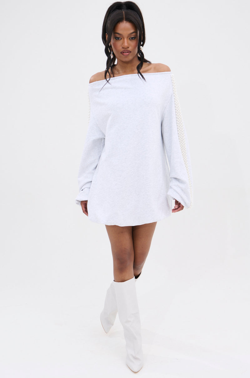 A woman models the GORGINA OFF THE SHOULDER SWEATSHIRT DRESS, featuring pearl-embellished sleeves, paired with white knee-high boots against a plain white background.