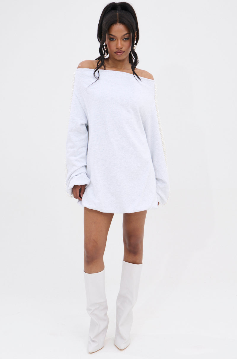 A woman with long, wavy dark hair looks slightly downward as she stands against a white background wearing the GORGINA OFF THE SHOULDER SWEATSHIRT DRESS with satin inner lining and knee-high white boots.