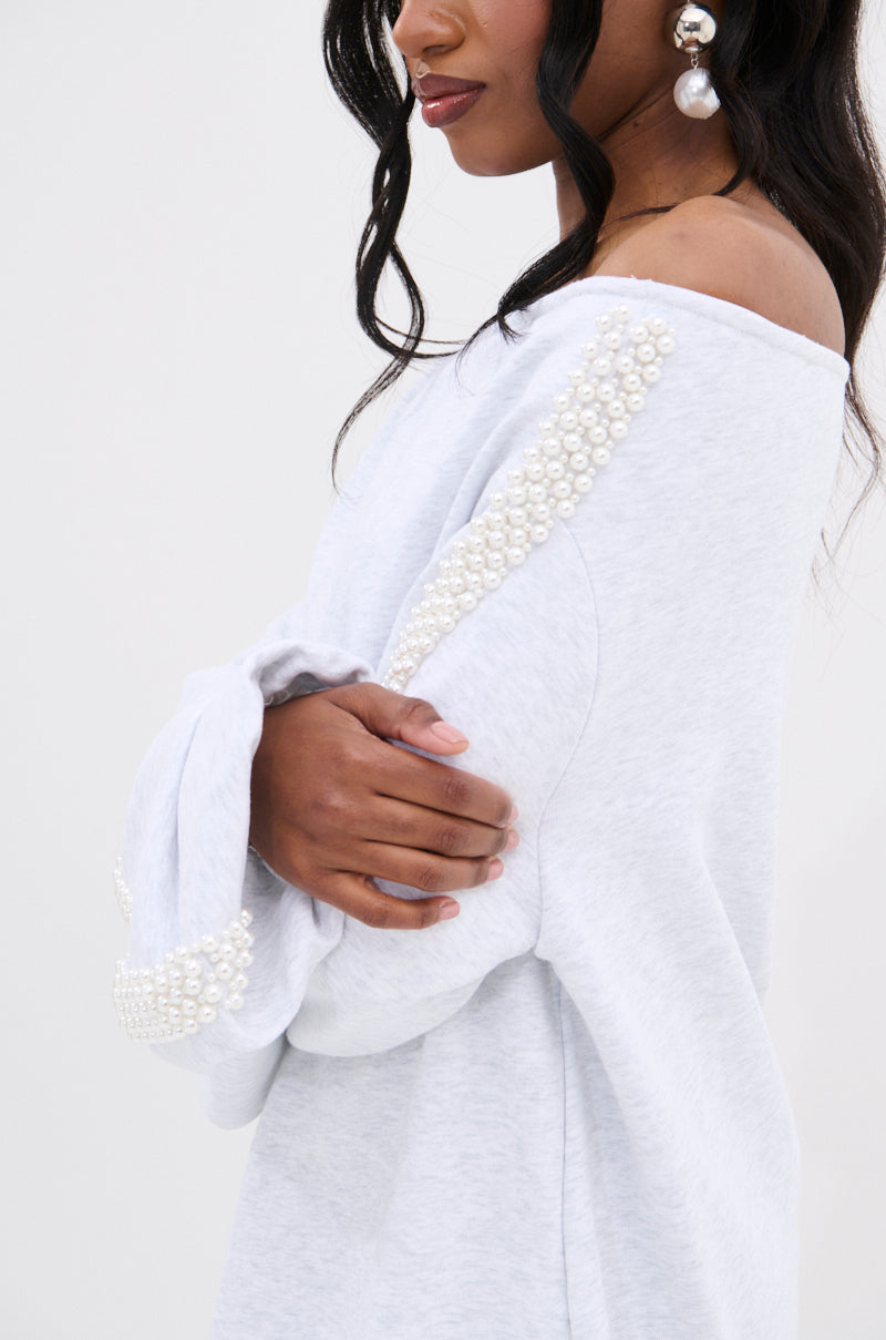 A woman with long dark hair wears the GORGINA OFF THE SHOULDER SWEATSHIRT DRESS in light gray, detailed with pearl embellishments on the sleeve and shoulder. She stands with arms crossed, completing the look with bold pearl earrings.