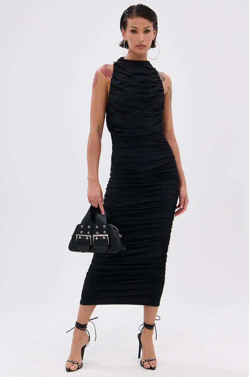 Wearing the BUSINESS IN FRONT PARTY IN BACK MIDI DRESS IN BLACK, a woman poses against a white background with a small black handbag, black strappy heels, slicked-back hair, and hoop earrings for a sleek, chic look.