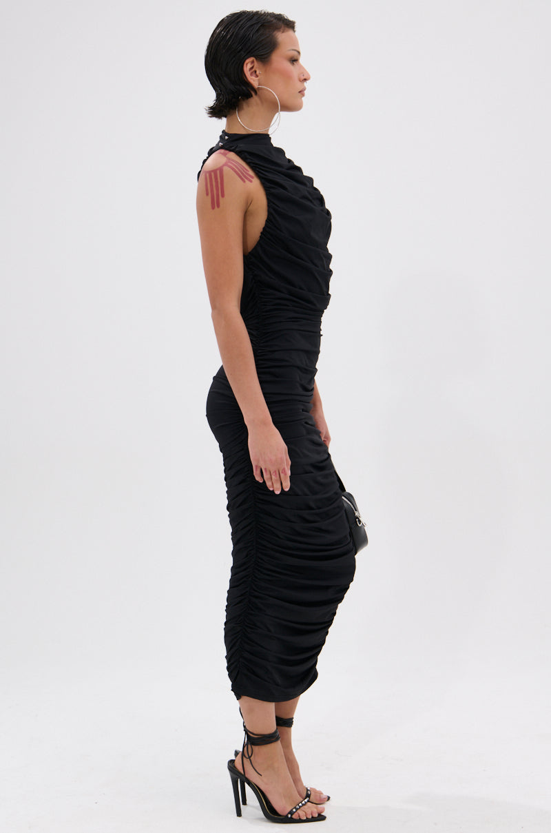 A person stands in profile against a plain background, wearing the BUSINESS IN FRONT PARTY IN BACK MIDI DRESS IN BLACK with black heeled sandals, holding a small bag. They have short, slicked-back hair and large hoop earrings.