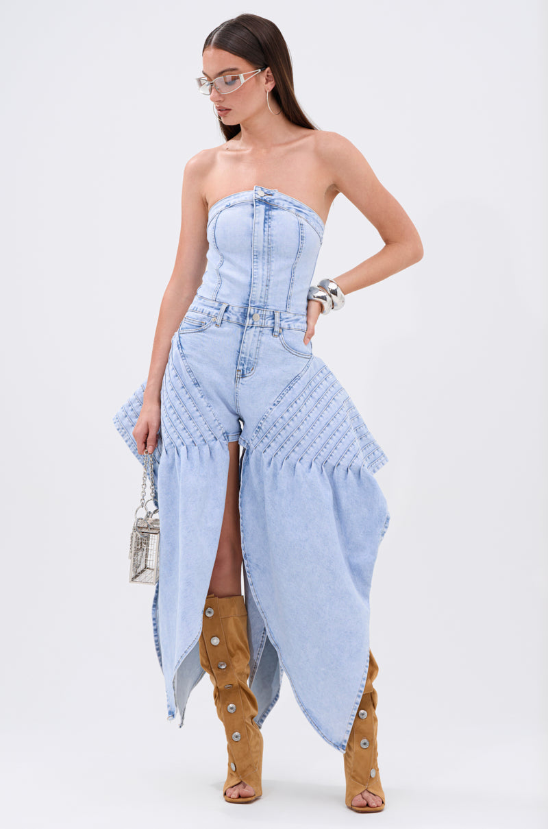 A woman wears the NO ONE LIKE ME DENIM ROMPER with tan boots, silver bangles, clear glasses, and carries a small textured handbag.
