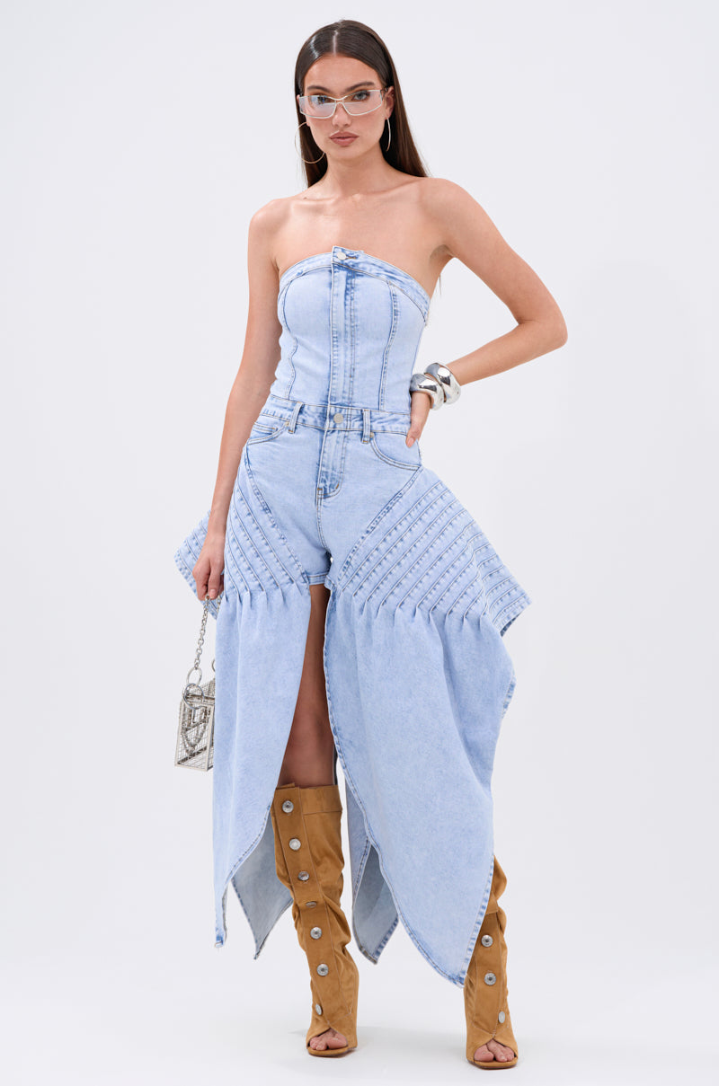 A woman wears the NO ONE LIKE ME DENIM ROMPER—a light blue, strapless, structured denim romper with a flared design and front slit. She accessorizes with clear glasses, tan knee-high boots, silver jewelry, and a small metallic bag.