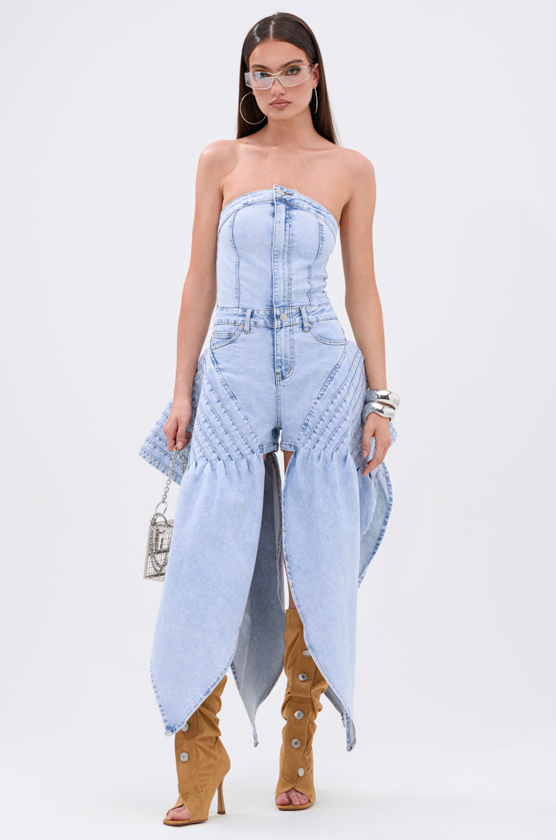 A woman showcases the NO ONE LIKE ME DENIM ROMPER, a strapless bustier light blue jumpsuit with wide pant legs, styled with tan knee-high open-toe boots, bold glasses, hoop earrings, bangles, and a small handbag for an avant-garde look.