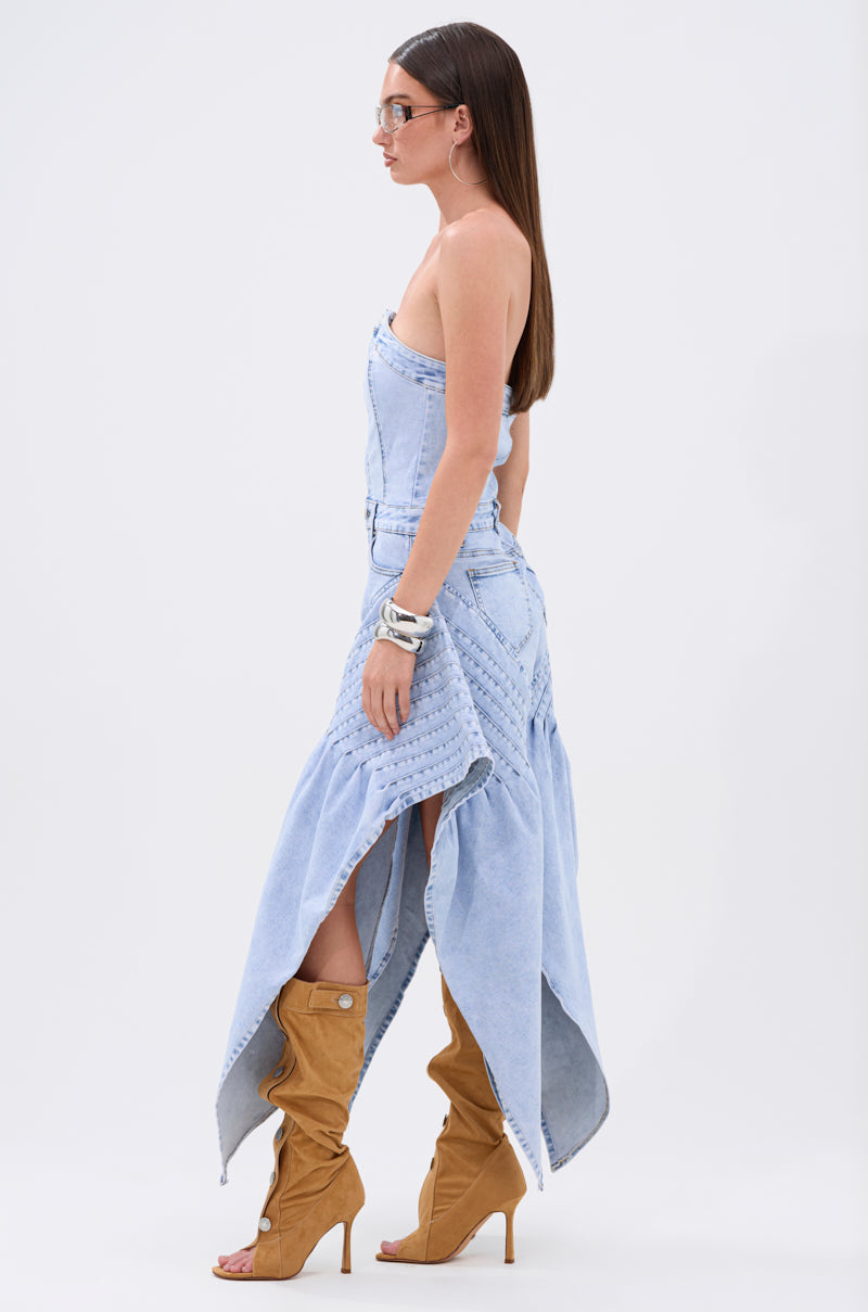 A woman with long straight hair wears the NO ONE LIKE ME DENIM ROMPER, featuring a strapless design and asymmetrical hem. She pairs it with tan heeled boots, silver bracelets, and glasses, standing in profile against a plain white background.